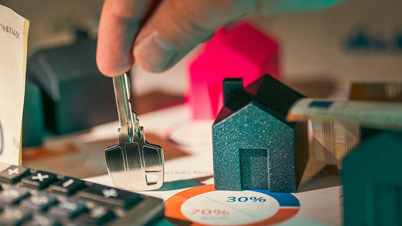 close up image of a hand placing a key on a table with house figures, calculator, and data reports about non-resident property mortgages