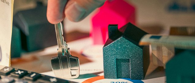 close up image of a hand placing a key on a table with house figures, calculator, and data reports about non-resident property mortgages