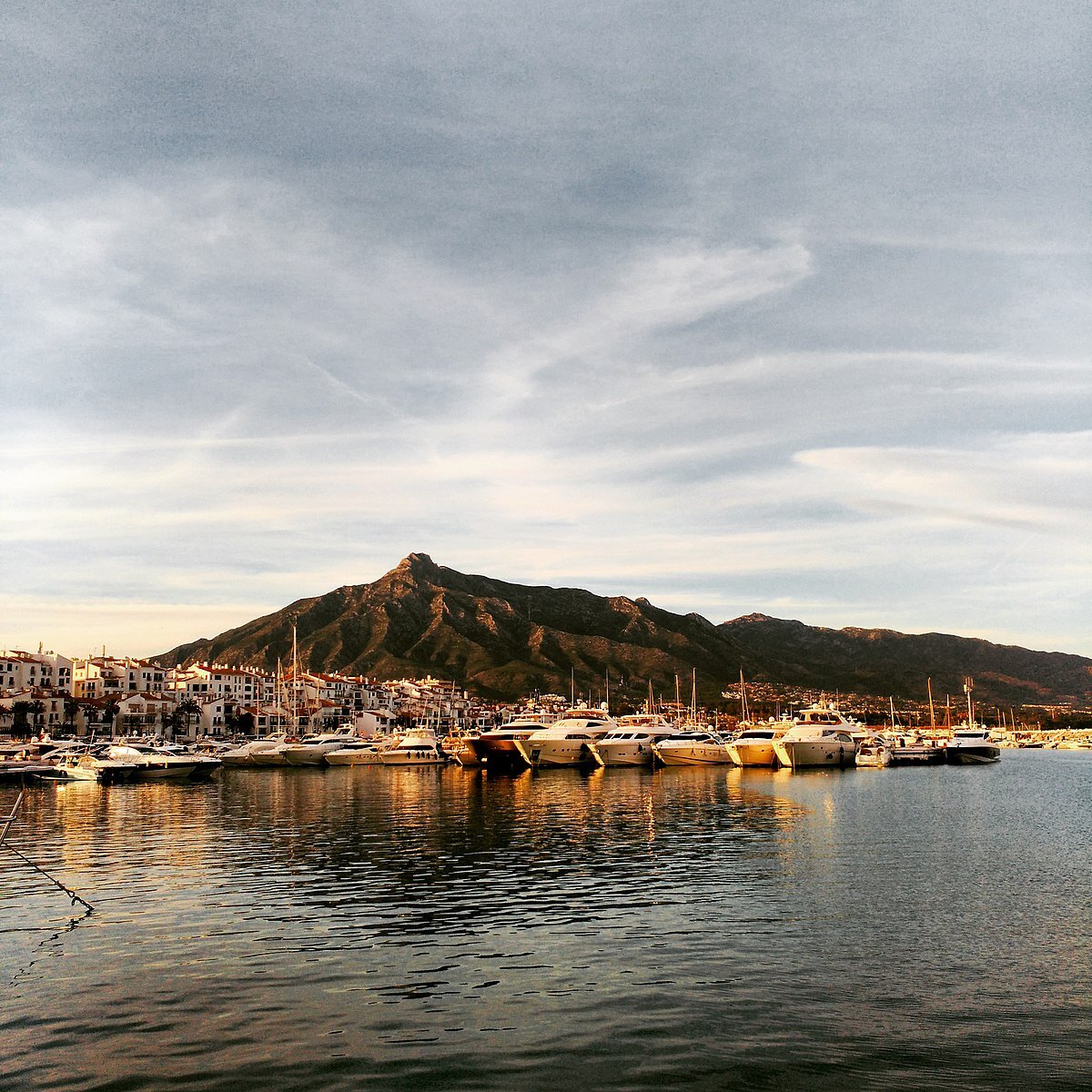 Sierra Blanca sedd från Puerto Banús.I bilden syns berg i bakgrunden och vatten i förgrunden. Några båtar ligger stilla på vattnet och bakom bergen går solen ner, vilket ger hela scenen ett lugnt och vackert intryck.