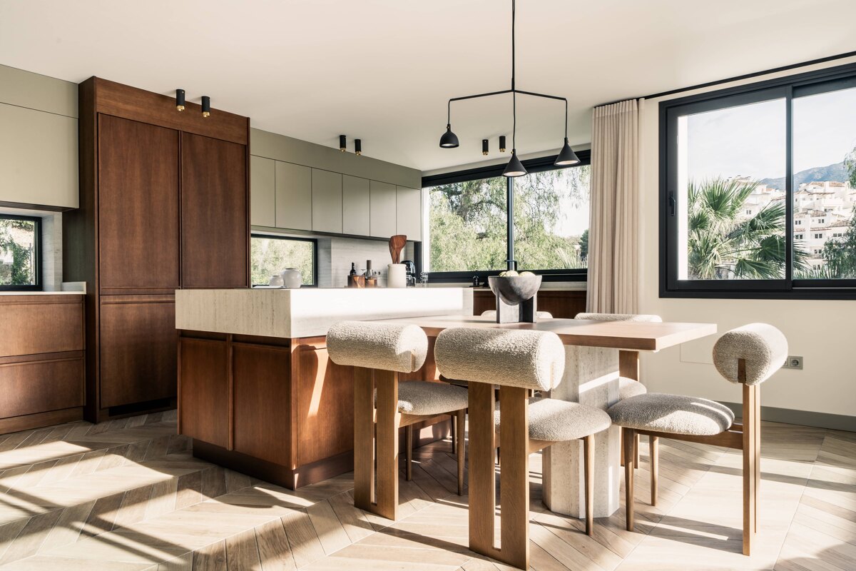 Villa Tira. Modern kitchen with wood cabinetry, neutral tones, a central island, and a dining area with upholstered chairs, framed by large windows with mountain views.