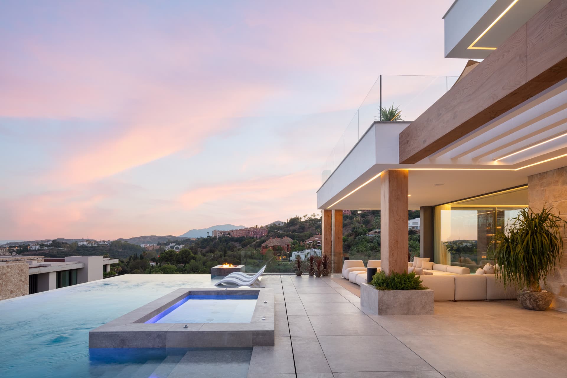 Modern villa terrace with an infinity pool, hot tub, and panoramic mountain views at sunset.