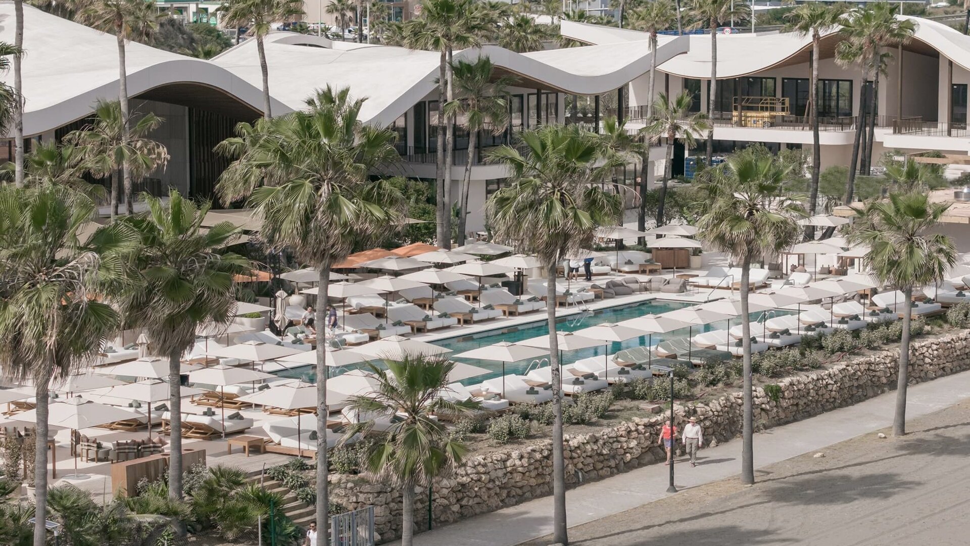 Aerial view of Sublim Beach Club in Estepona, showcasing its sleek architecture, elegant poolside loungers, and curated parasols, surrounded by lush palm trees and a scenic beachfront promenade.