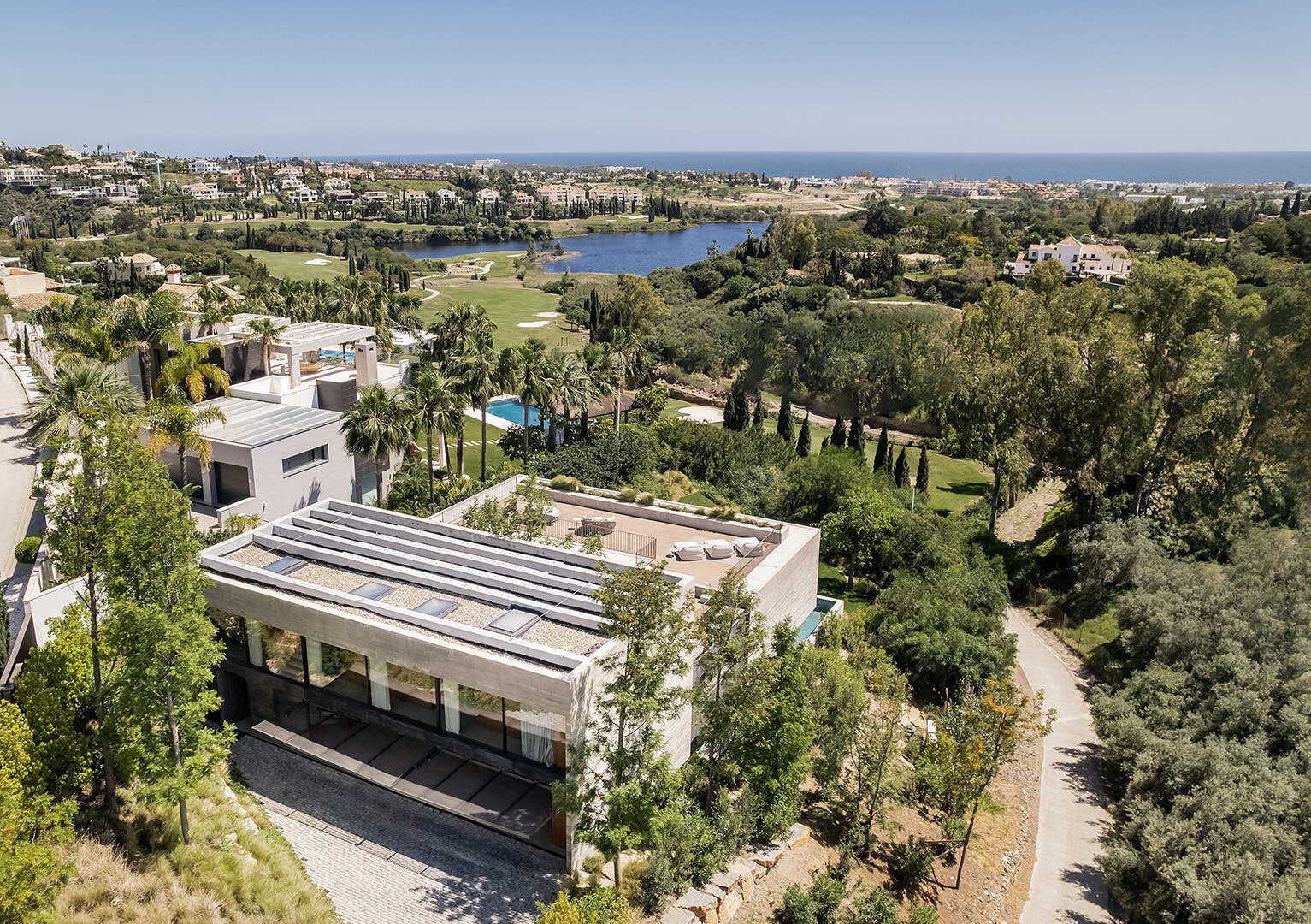 Aerial view of Los Flamingos, showcasing a contemporary villa surrounded by lush greenery, rolling golf courses, and a scenic lake, with the Mediterranean Sea in the background.