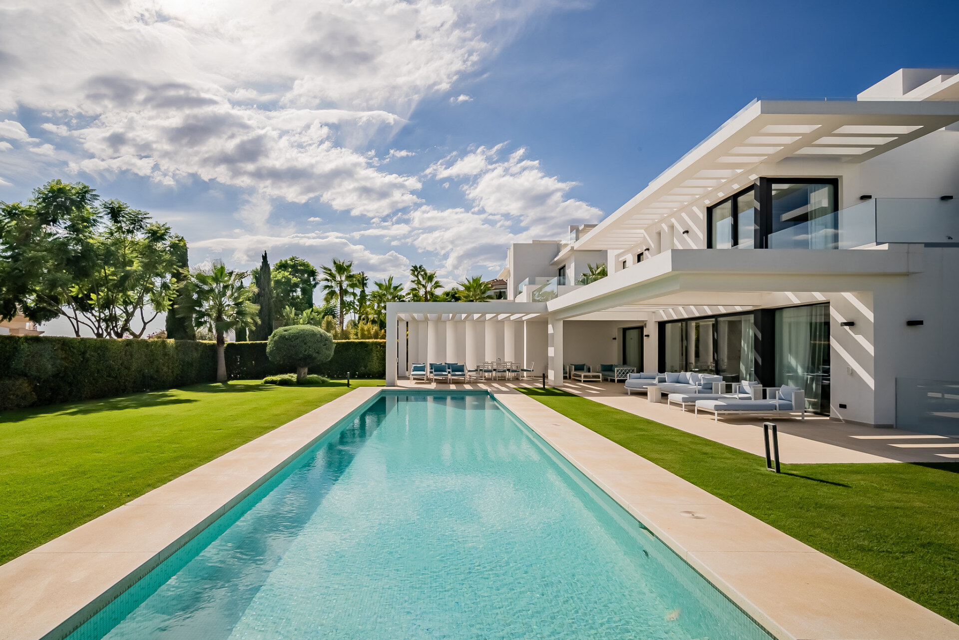 Modern white villa with clean architectural lines, a large swimming pool, and lush gardens, set against a backdrop of palm trees and a blue sky.