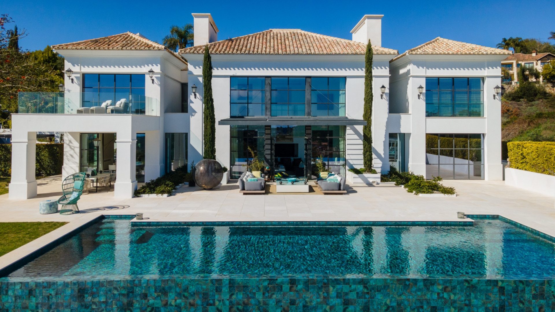 Villa Jasmine in Los Flamingos, a striking white villa with expansive glass windows, terracotta roof tiles, and an elegant outdoor area featuring a designer infinity pool, manicured landscaping, and stylish lounge spaces.