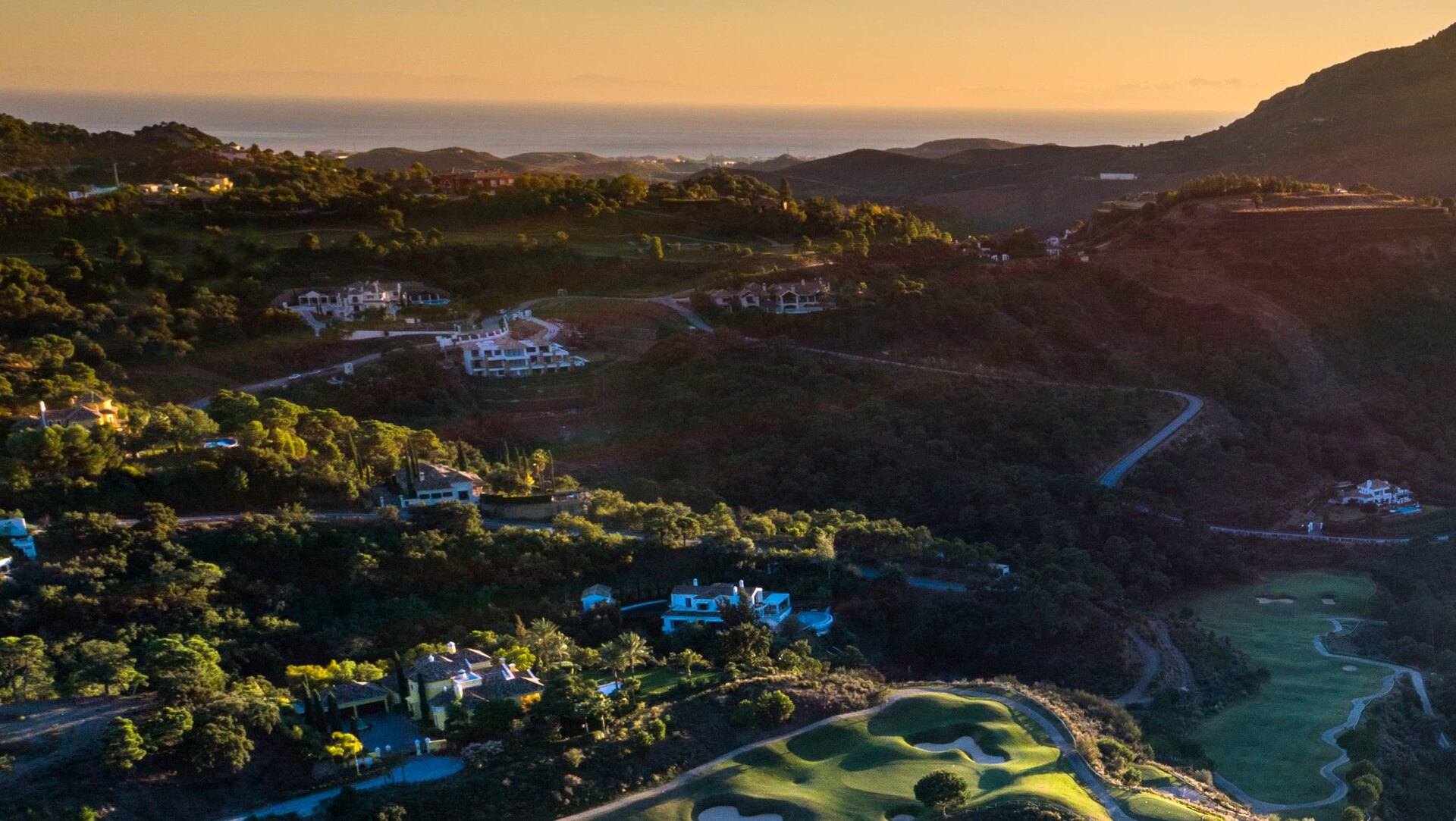 Aerial view of La Zagaleta at sunset, showcasing rolling hills, luxury villas, winding roads, and a golf course with the Mediterranean Sea in the background.