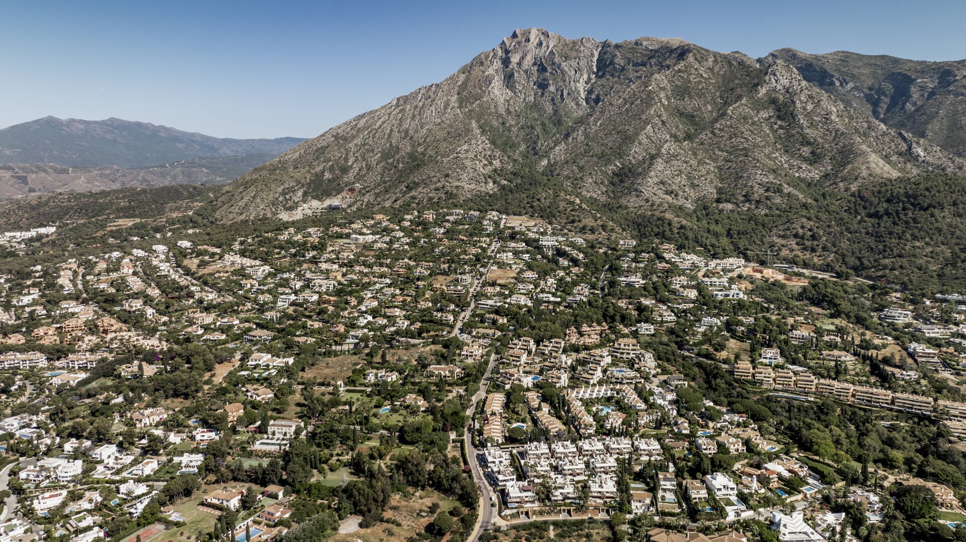 Aerial view of the Sierra Blanca area, featuring a landscape of luxurious villas surrounded by lush green hills, with the dramatic backdrop of the towering mountain range.