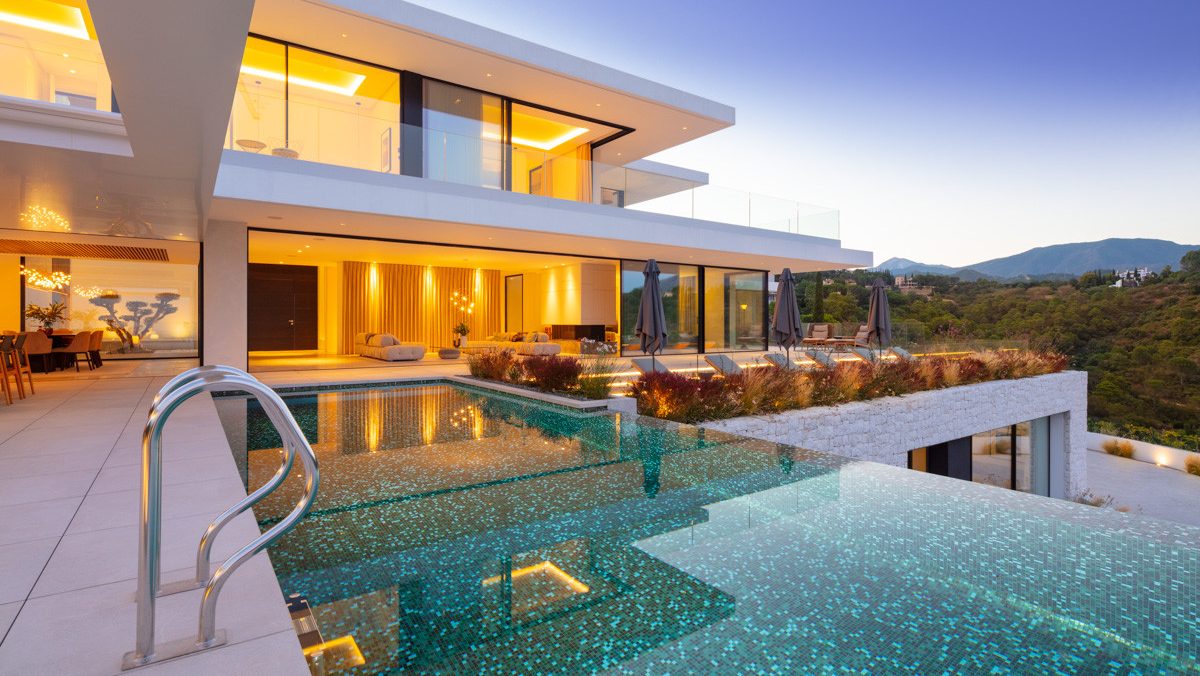 Modern villa in El Madroñal with sleek architecture, floor-to-ceiling windows, and an infinity pool overlooking lush hills at dusk.