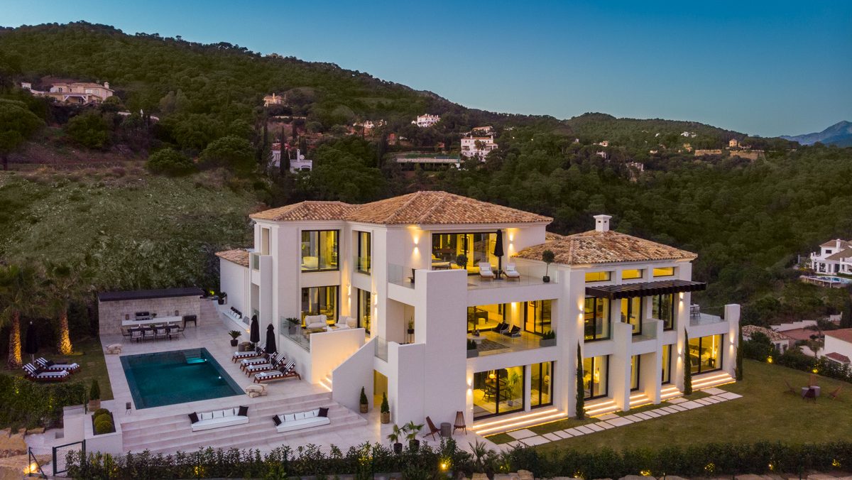 Elegant villa in El Madroñal with a classic-modern design, large terraces, an illuminated pool, and panoramic mountain views at dusk.