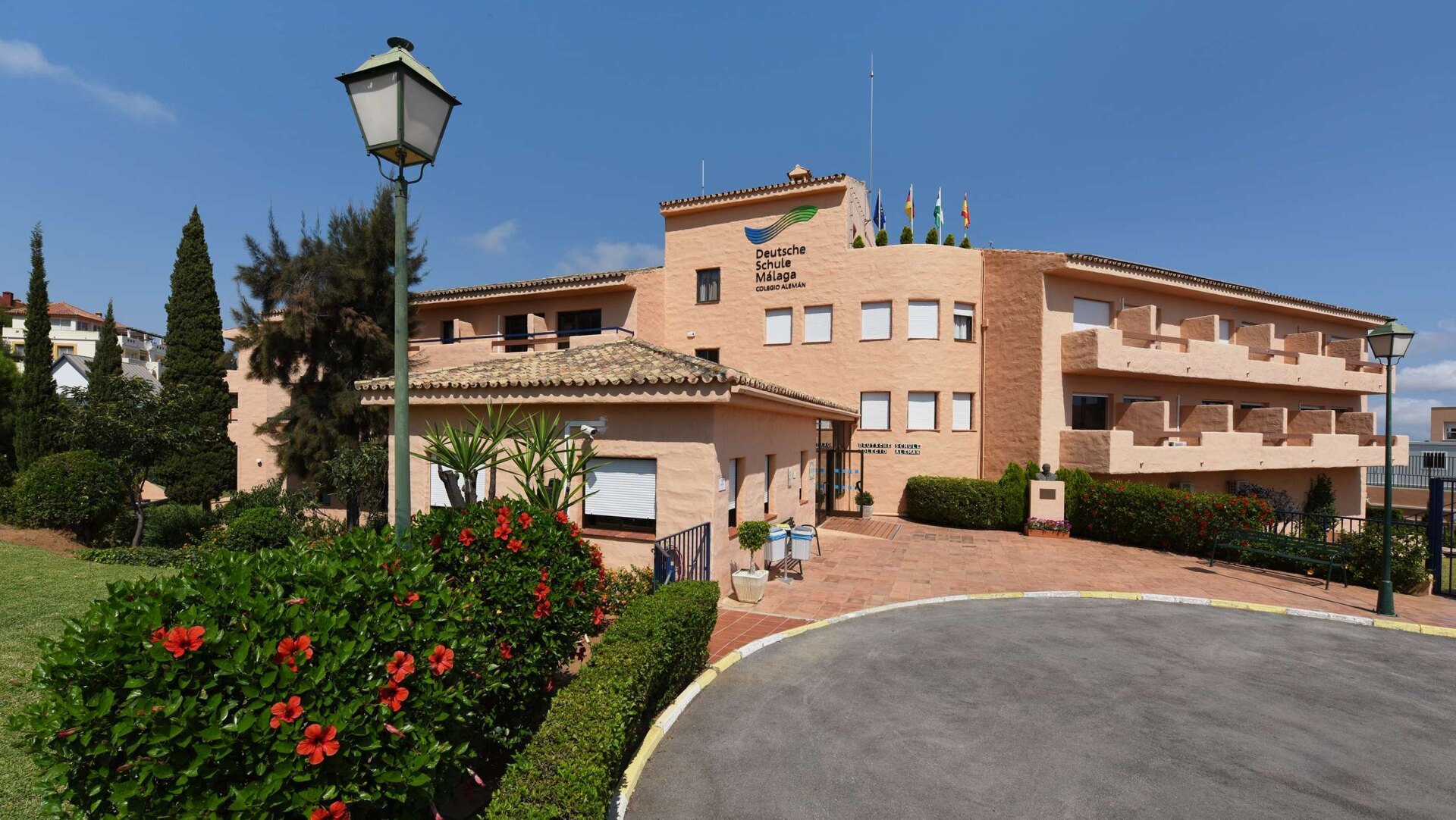 Deutsche Schule Málaga entrance with terracotta buildings, lush greenery, and red hibiscus flowers under a clear blue sky.