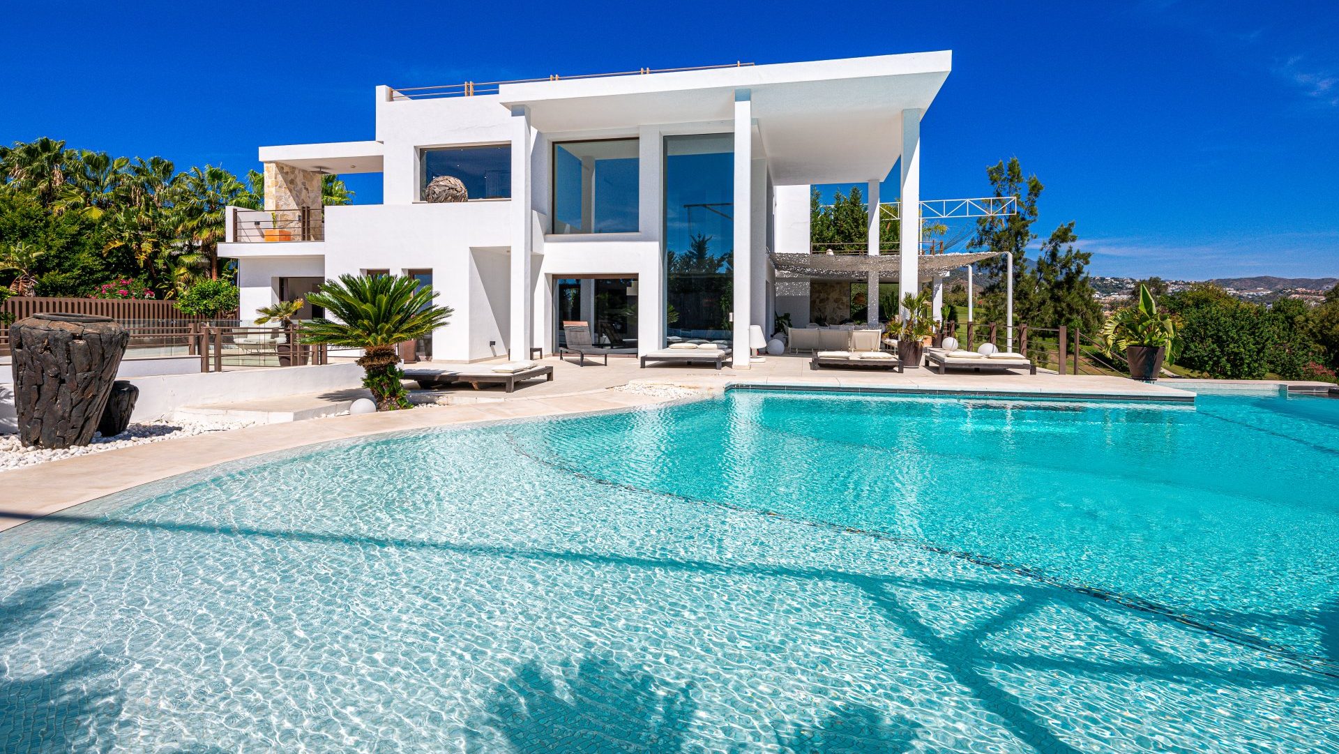 Contemporary white villa in Benahavís with floor-to-ceiling windows, expansive terraces, and a large turquoise pool surrounded by lush greenery.