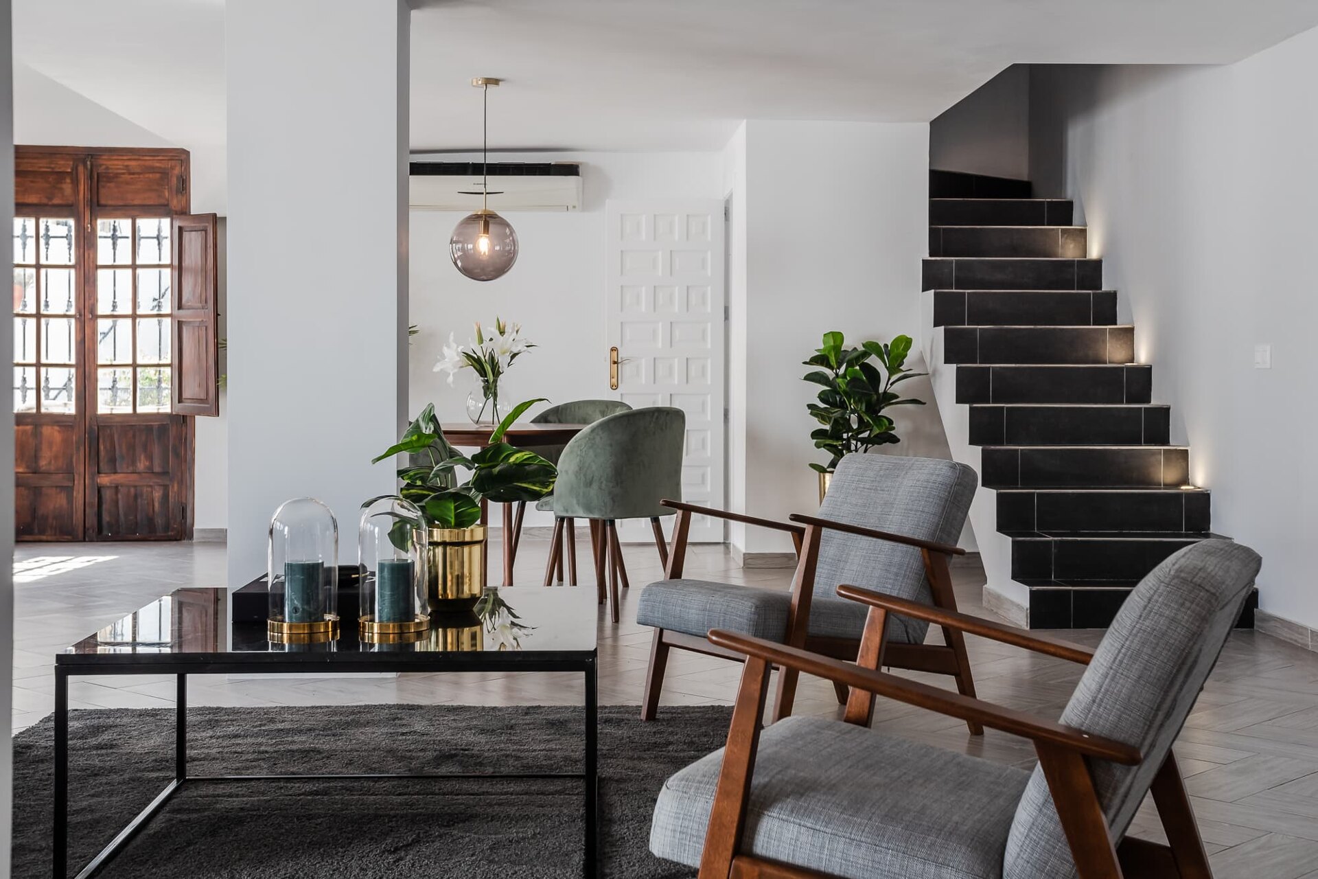 Modern living space with wooden accents, grey armchairs, a black staircase with LED lighting, and a dining area with green velvet chairs.