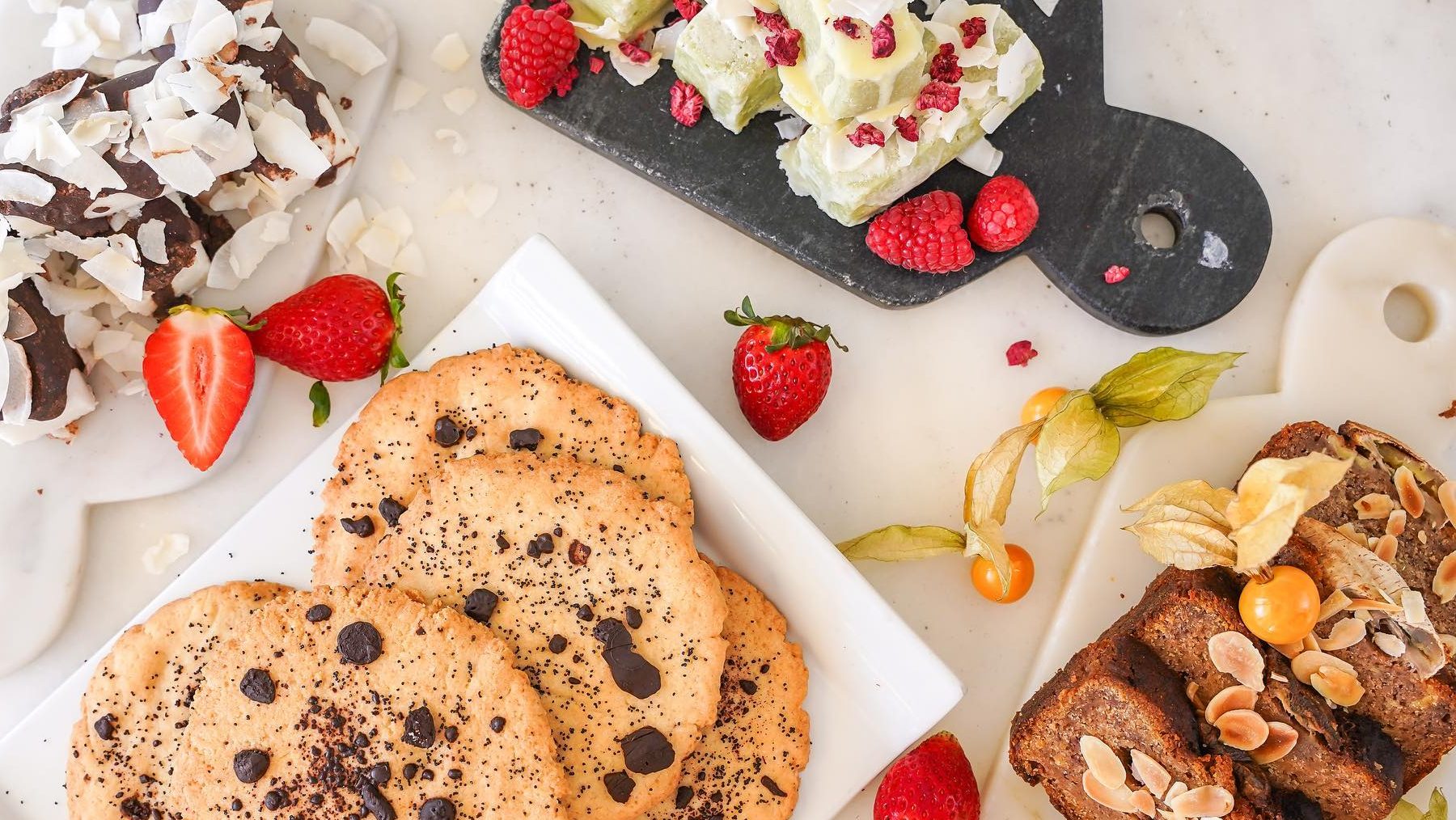 Assorted desserts on a marble table, including chocolate chip cookies, toasted bread with almonds, and fruit-topped treats with strawberries and raspberries.