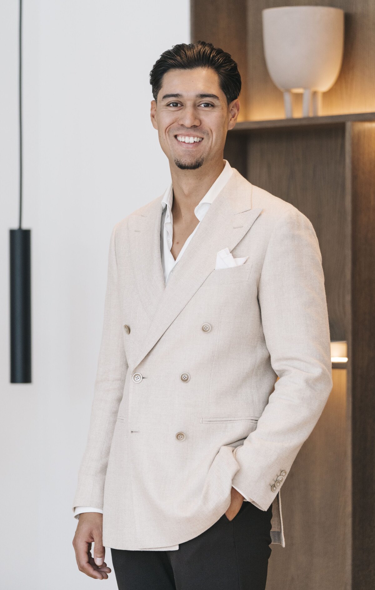 A stylish man in a beige double-breasted blazer, smiling confidently in a modern interior setting.