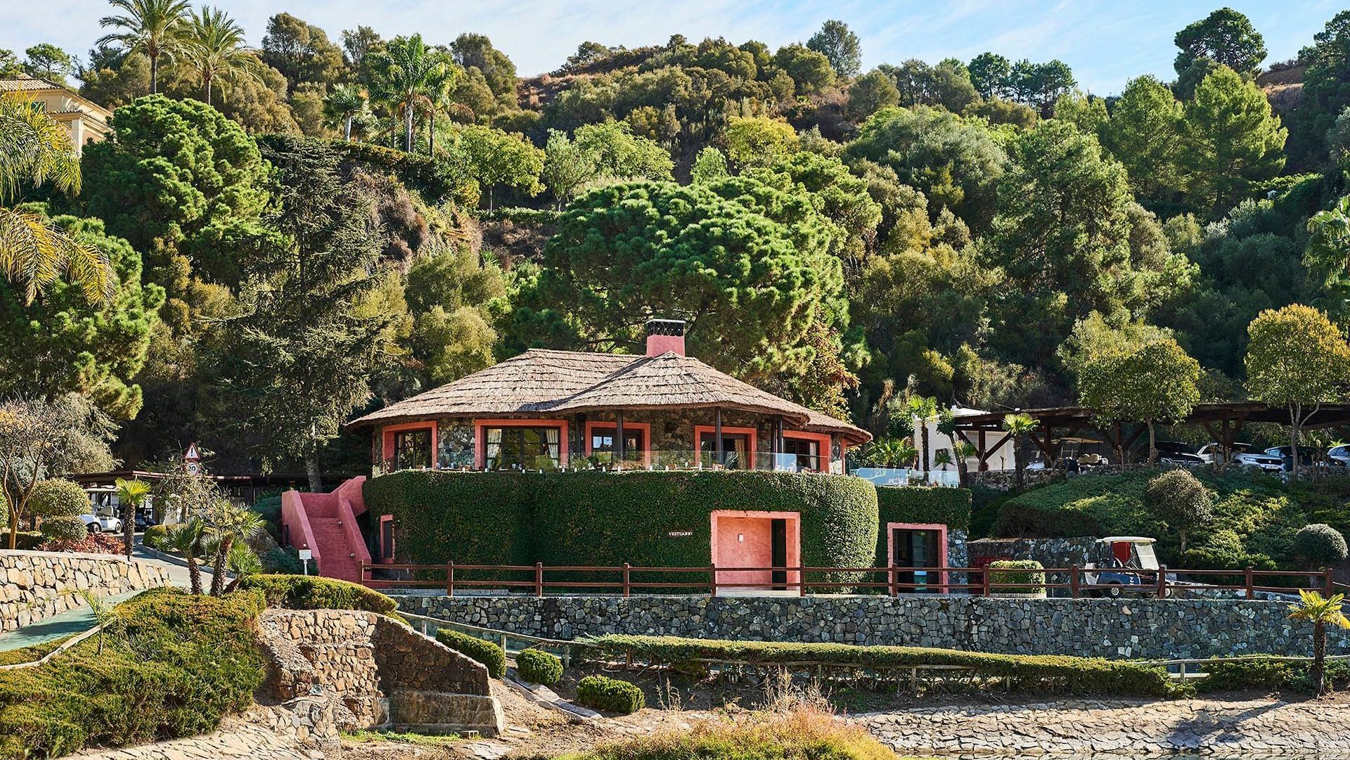 Rustic restaurant on a golf course in Marbella, featuring a circular design, red accents, and lush greenery, set against a hillside landscape.