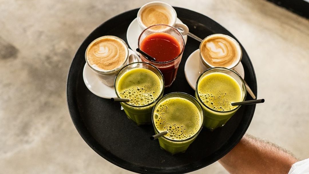 A tray with a selection of drinks, including cups of coffee with latte art, green juices, and a glass of fresh orange juice, arranged neatly on a black tray.