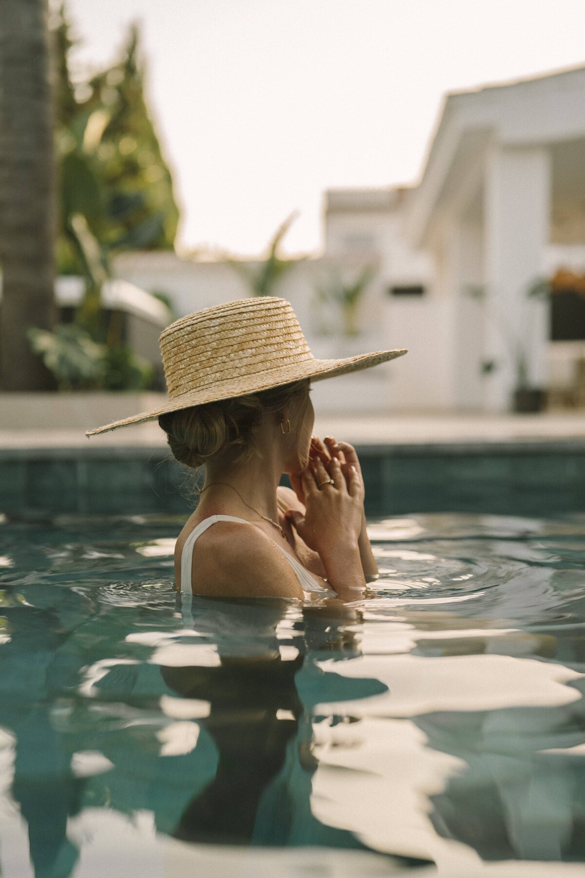 Villa María. Woman in a straw hat relaxing in a Marbella villa pool, surrounded by lush greenery and elegant white architecture.