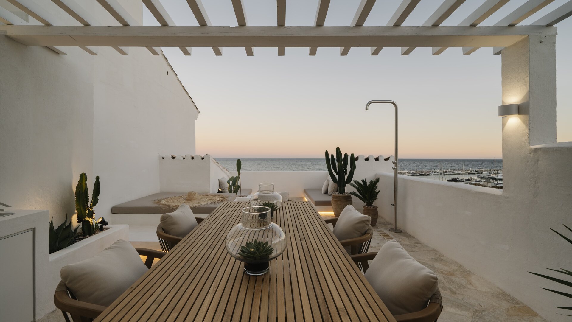 A stylish terrace with a wooden dining table, cactus plants, and a view of Puerto Banús marina at sunset, featuring a warm, tranquil ambiance.