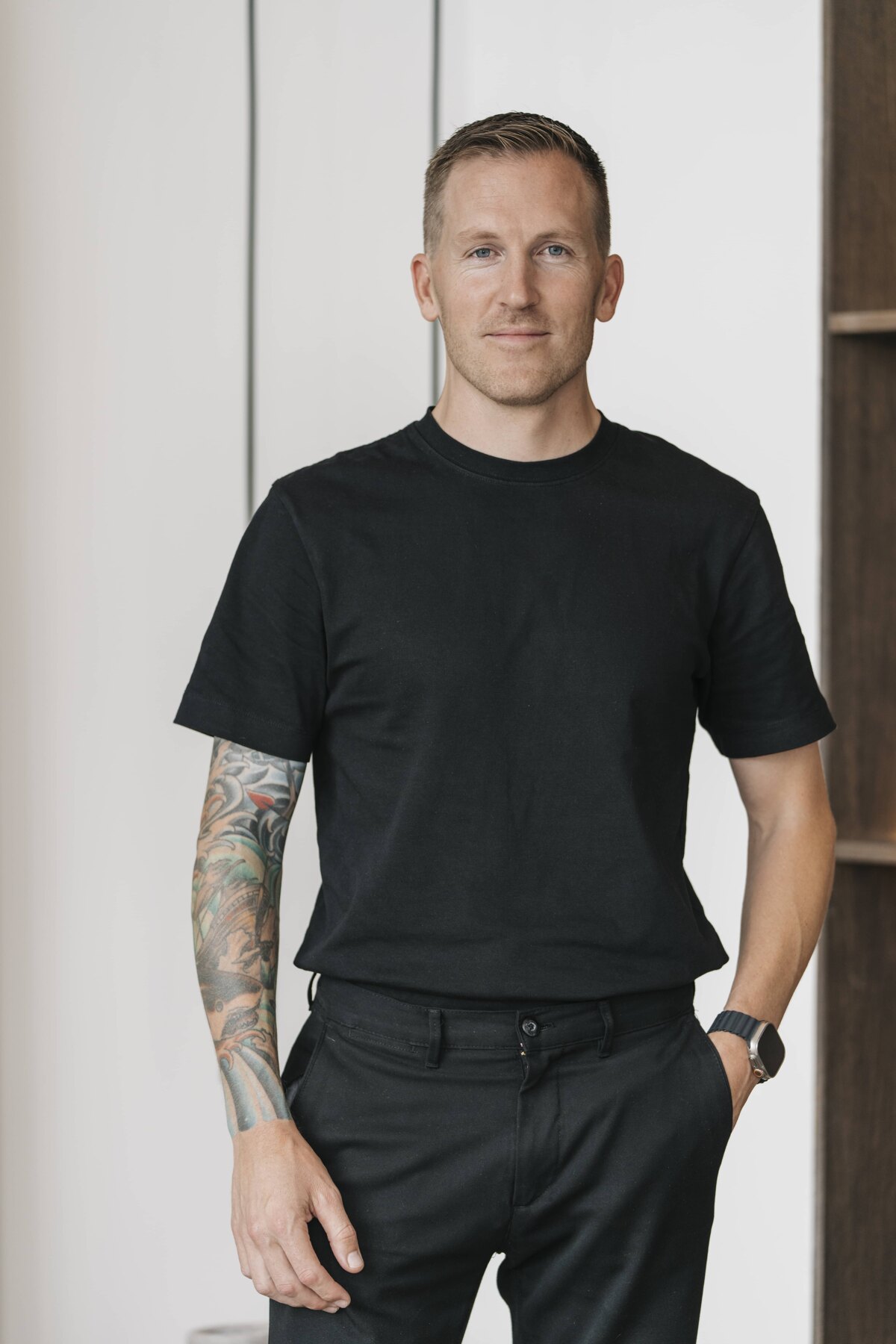 A man with short light brown hair and a tattoo on his arm, wearing a black t-shirt and black pants. He is standing in front of a bookshelf with a relaxed posture, hands in his pockets.