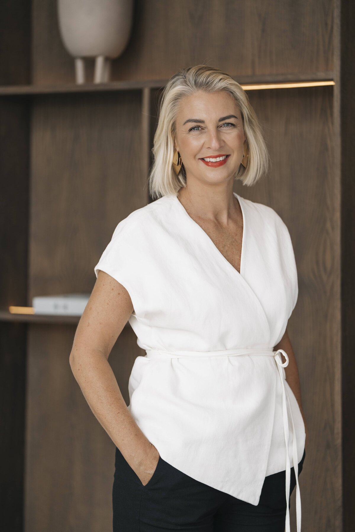 A woman with short platinum blonde hair and blue eyes, wearing a white blouse and black pants. She is standing with one hand in her pocket and smiling in front of a wooden bookshelf.