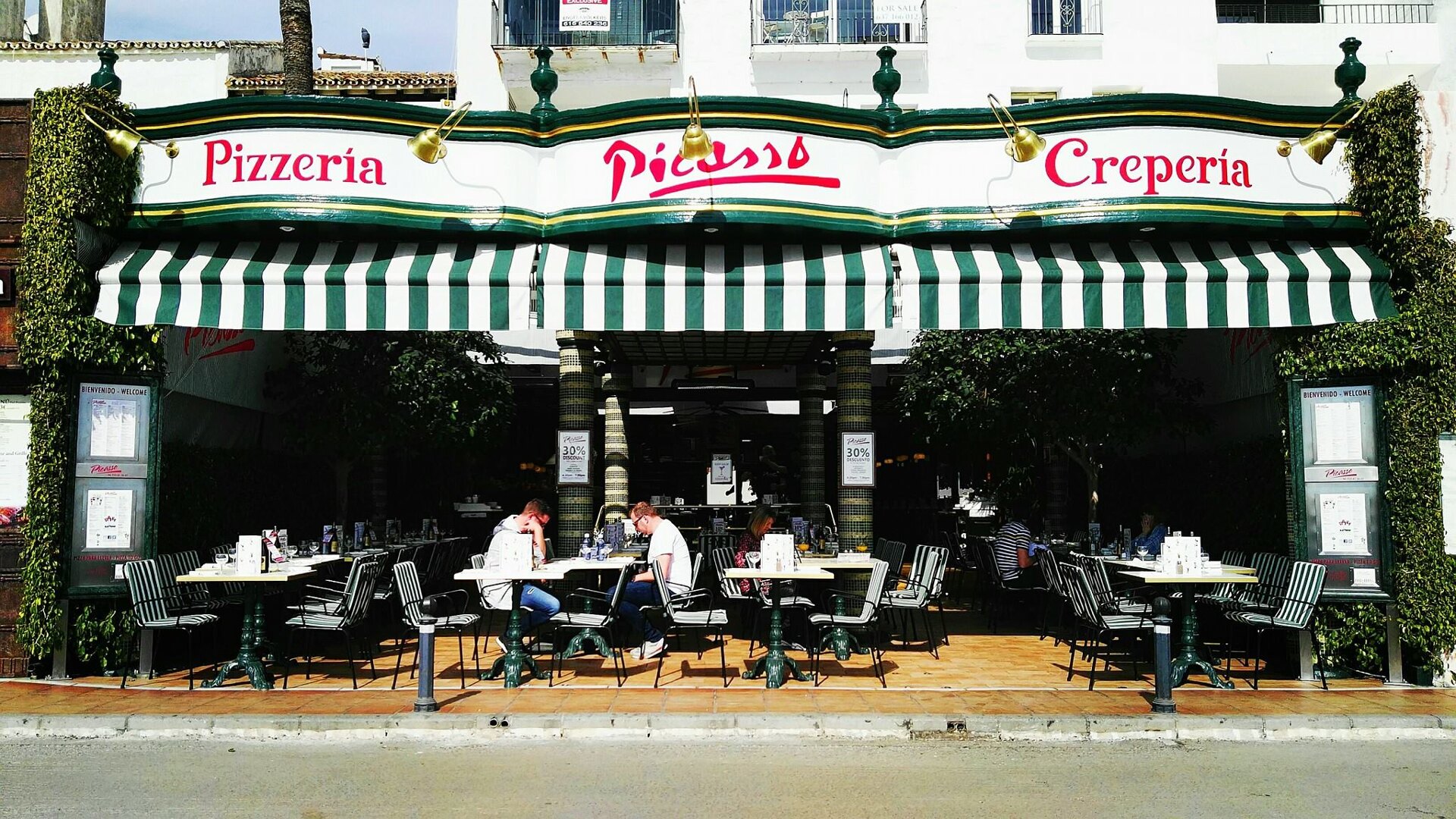 The entrance to the Picasso Pizzeria and Crepería, with green and white striped awnings, offering outdoor seating in a lively atmosphere in Puerto Banús. Guests are seated at tables with a welcoming sign in front.