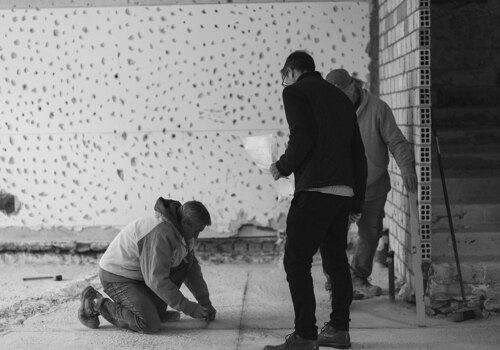 Black and white image of three men working on a construction site, measuring and discussing renovation plans in an unfinished space.