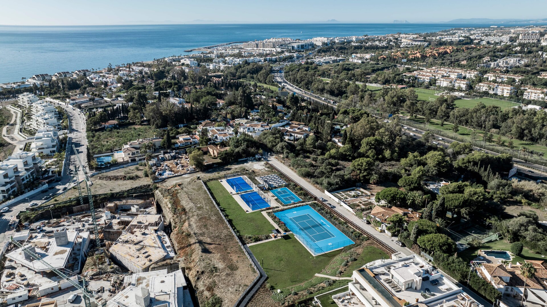 Aerial view of a coastal area with modern white villas, tennis and padel courts, lush greenery, and the sea stretching to the horizon.