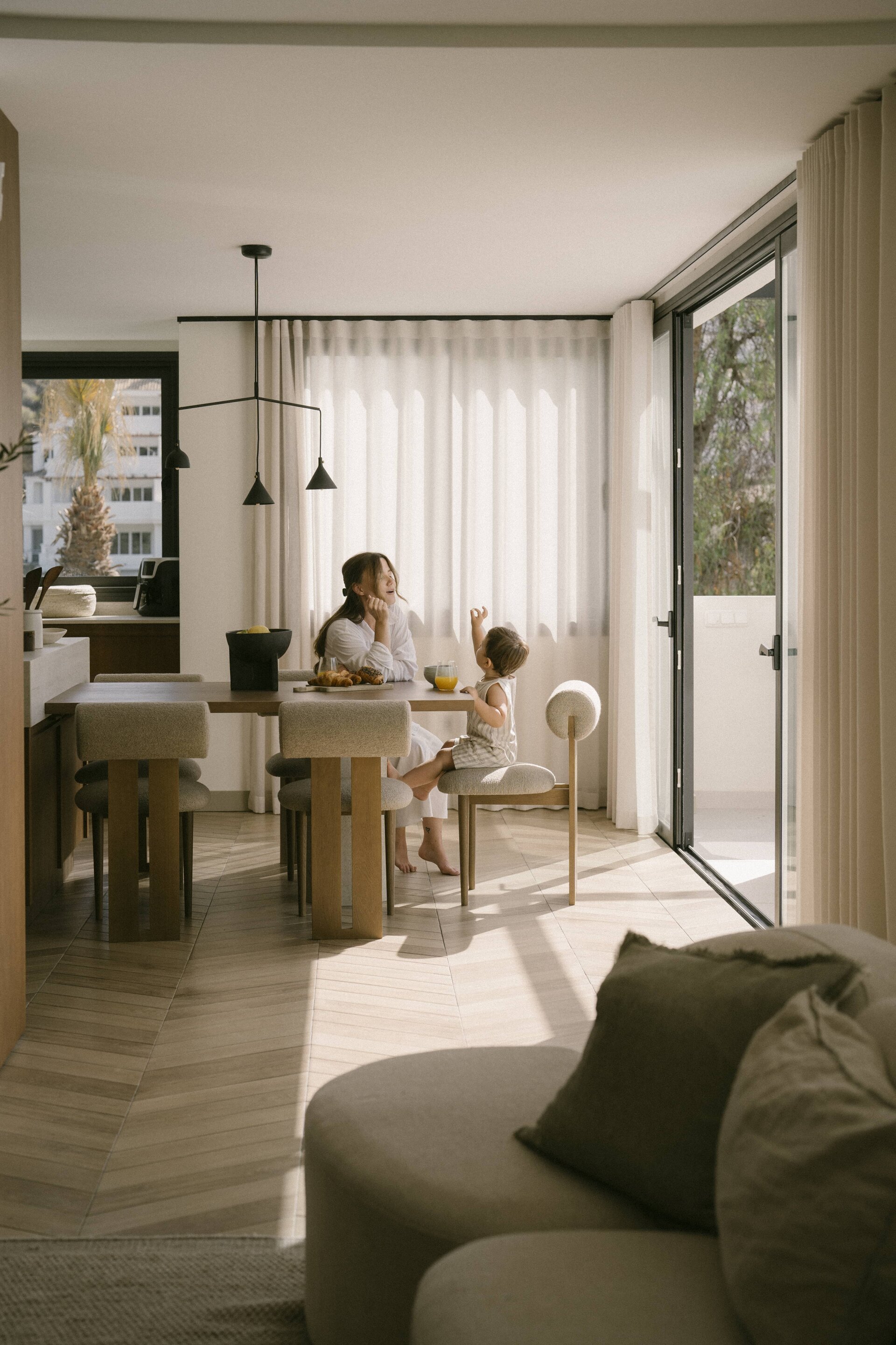 Mother and child in a sunlit dining area with wooden floors, neutral decor, and floor-to-ceiling curtains, next to an open terrace.