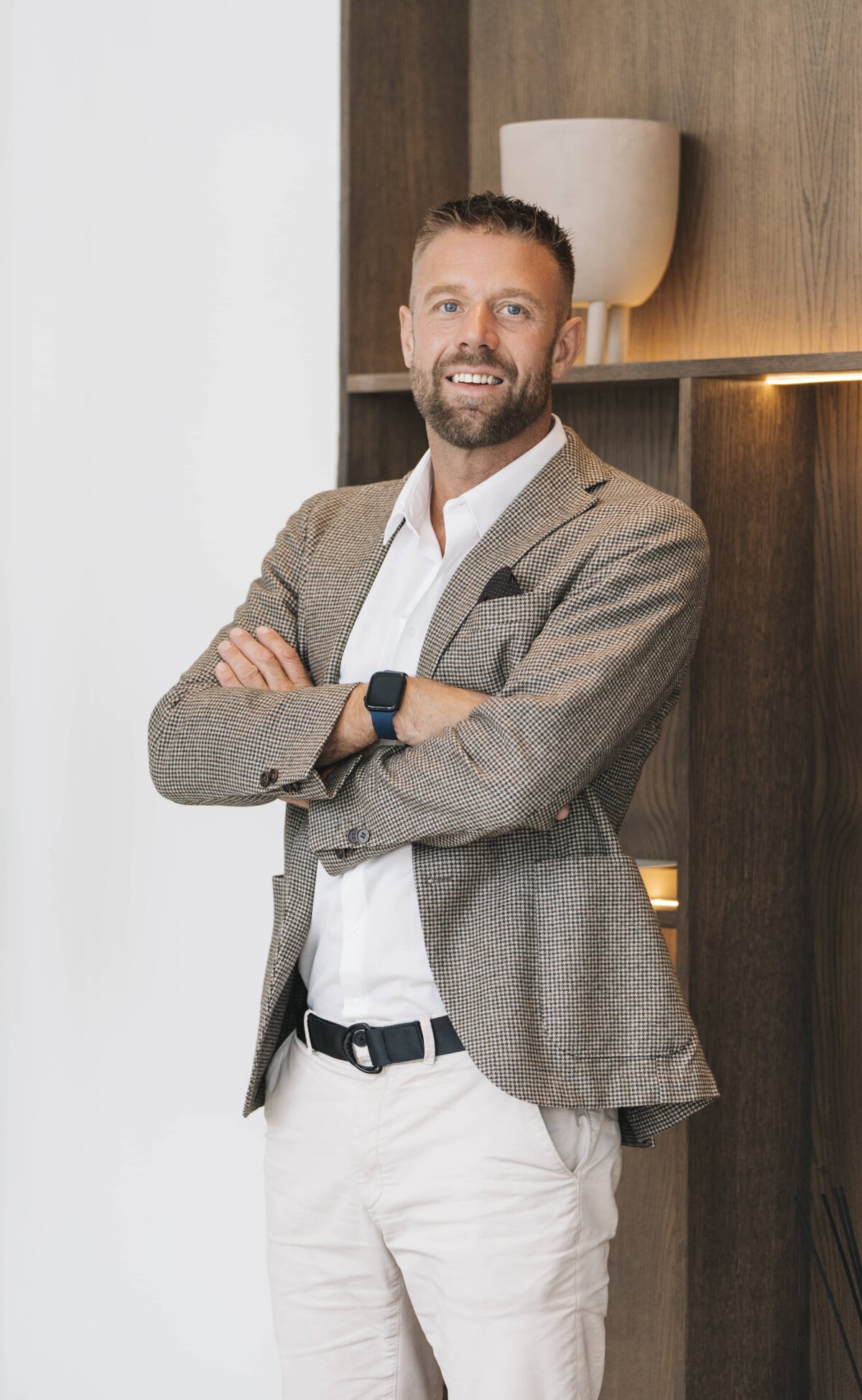 Andreas Grenthe: Male, fair skin with light brown eyes, short light brown hair. He is dressed in a beige blazer over a white shirt, paired with beige trousers. He smiles confidently with his arms crossed in front of a modern background featuring warm lighting and minimalist decor.