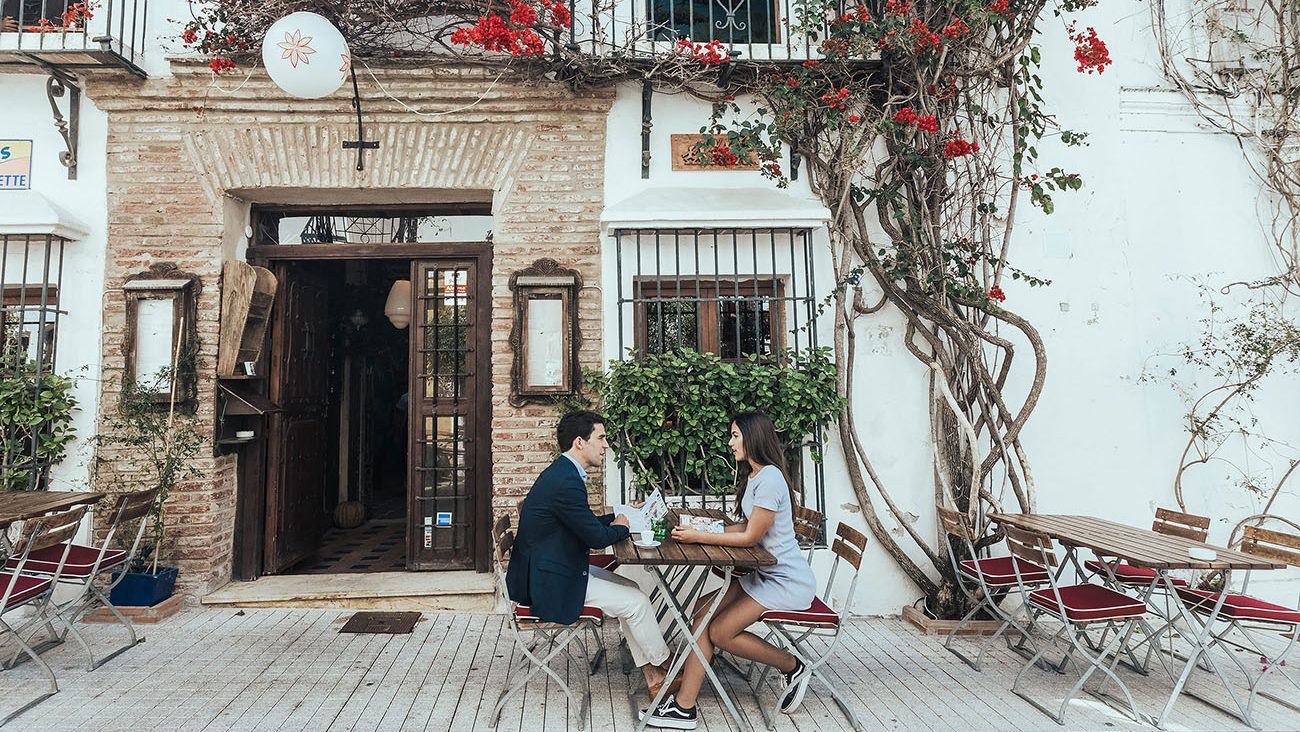 A young man and woman enjoying a drink at a table on a street in Marbella Old Town, surrounded by vibrant fuchsia bougainvillea flowers.