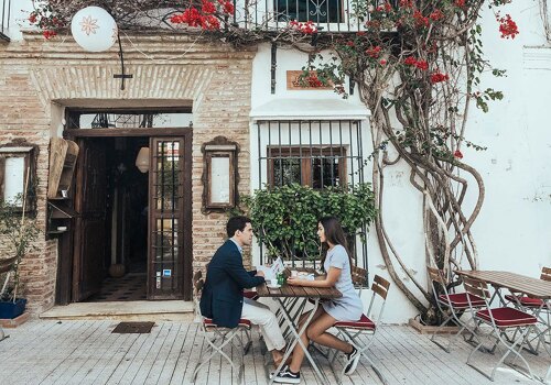 A young man and woman enjoying a drink at a table on a street in Marbella Old Town, surrounded by vibrant fuchsia bougainvillea flowers.