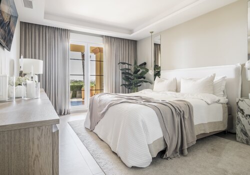 Elegant bedroom in Cumbres Apartment, featuring a soft neutral palette, plush bedding, a marble side table, and floor-to-ceiling curtains leading to a private terrace with sea views.