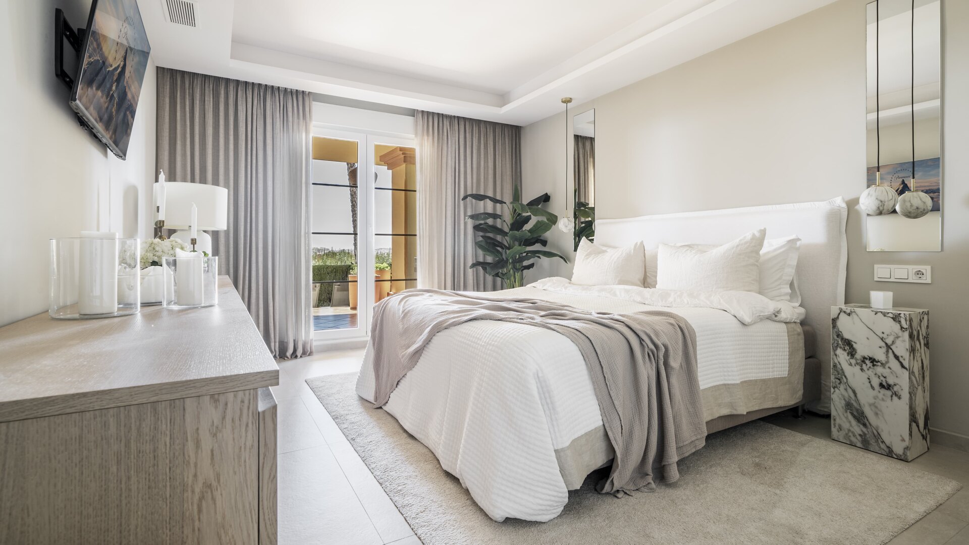 Elegant bedroom in Cumbres Apartment, featuring a soft neutral palette, plush bedding, a marble side table, and floor-to-ceiling curtains leading to a private terrace with sea views.