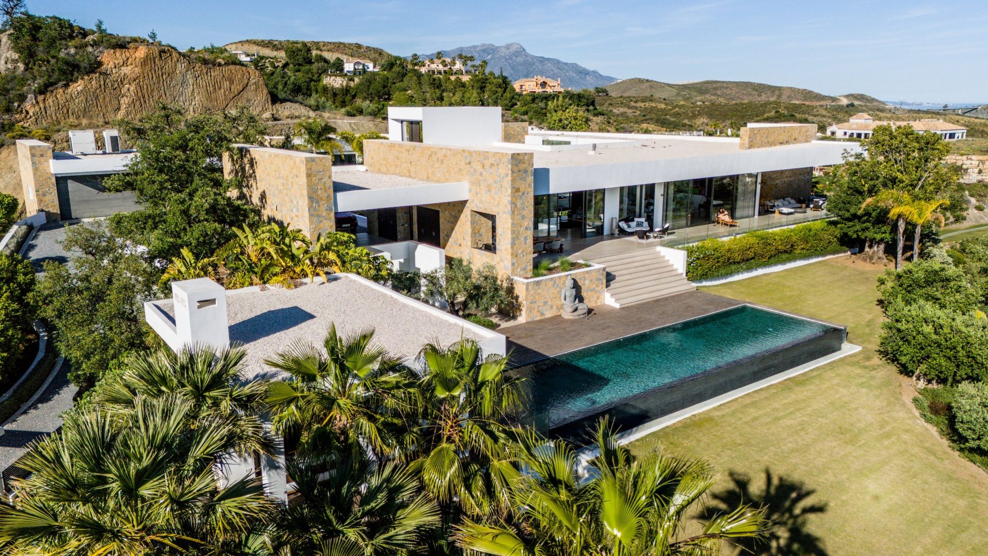 Modern villa in Marbella with a minimalist design, stone and glass façade, infinity pool, and panoramic mountain views, surrounded by lush greenery.