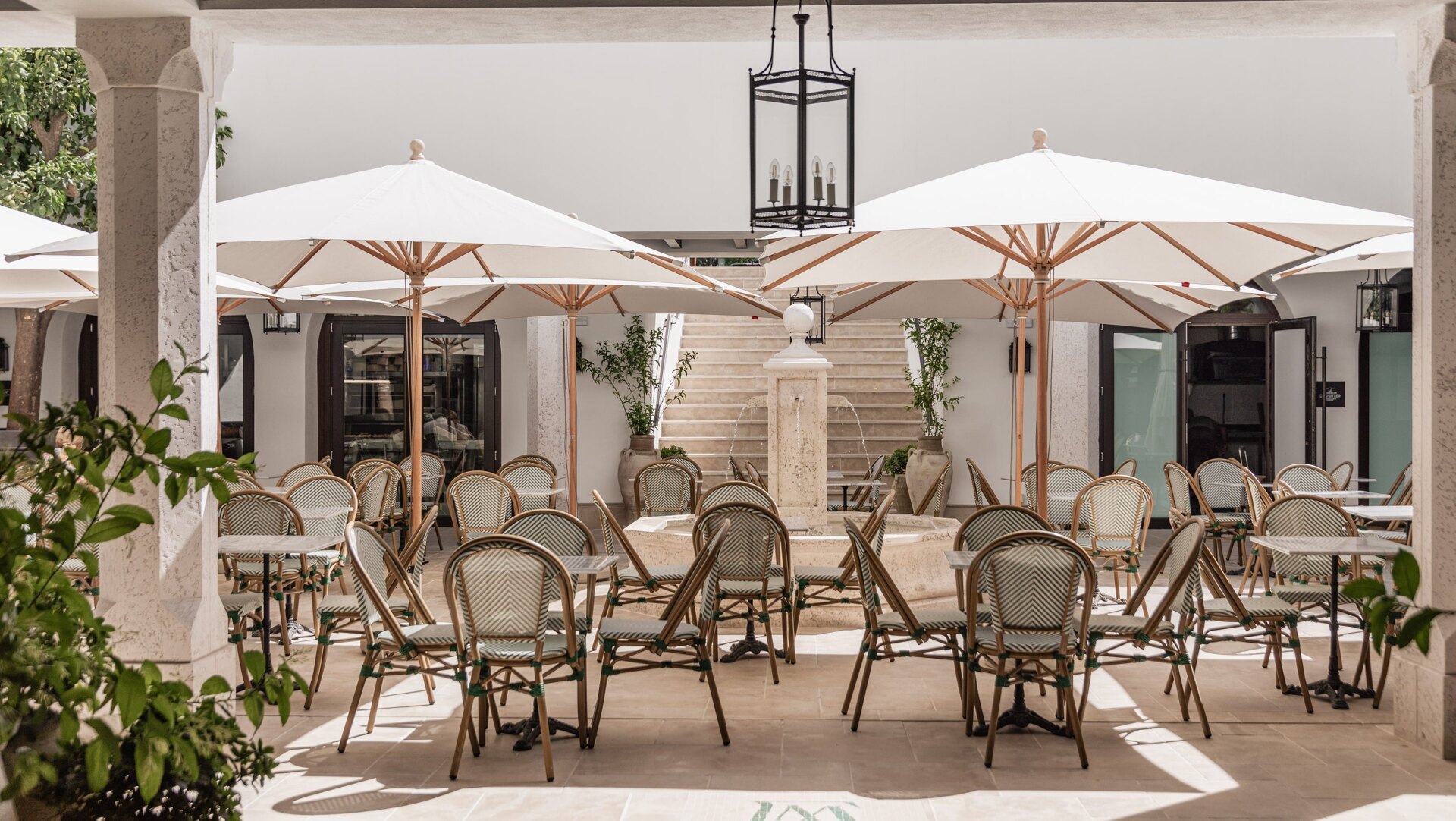 Elegant courtyard with woven chairs, white umbrellas, and a central stone fountain, framed by arched doors and lush greenery.