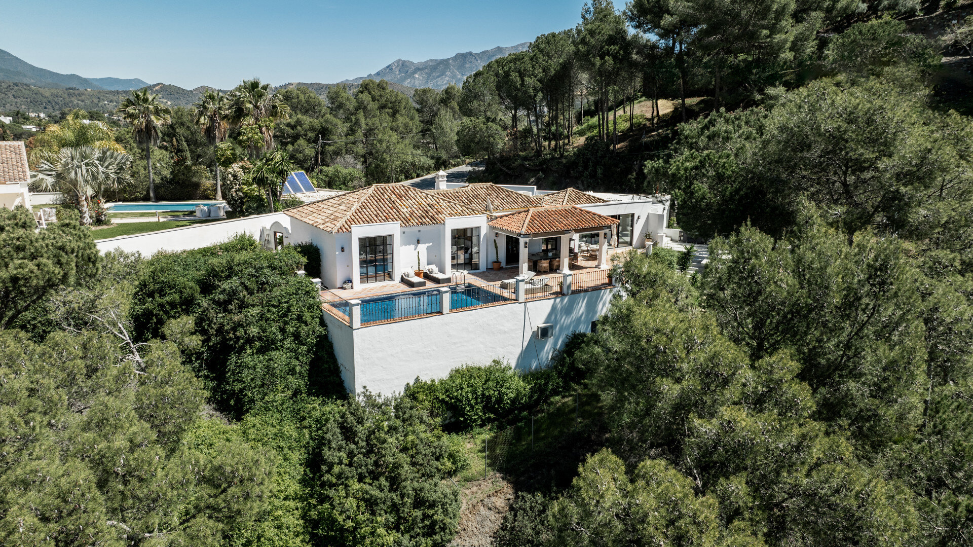 Aerial view of a white villa surrounded by lush greenery and pine trees, featuring a private pool and a shaded terrace with outdoor seating, set against a mountainous backdrop.