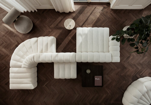 An aerial view of a luxurious living room featuring a unique, curved sofa by NORR11 in soft beige fabric. The sofa, with its distinctive segmented design, wraps around the room and is complemented by a matching lounge chair. A dark wooden coffee table sits at the center, with a book titled 