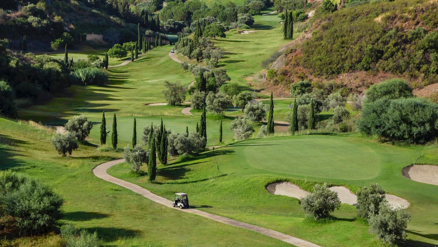 Rolling fairways and manicured greens of Villa Padierna Golf Club, framed by cypress trees and Mediterranean vegetation, with a winding path leading through the lush landscape.