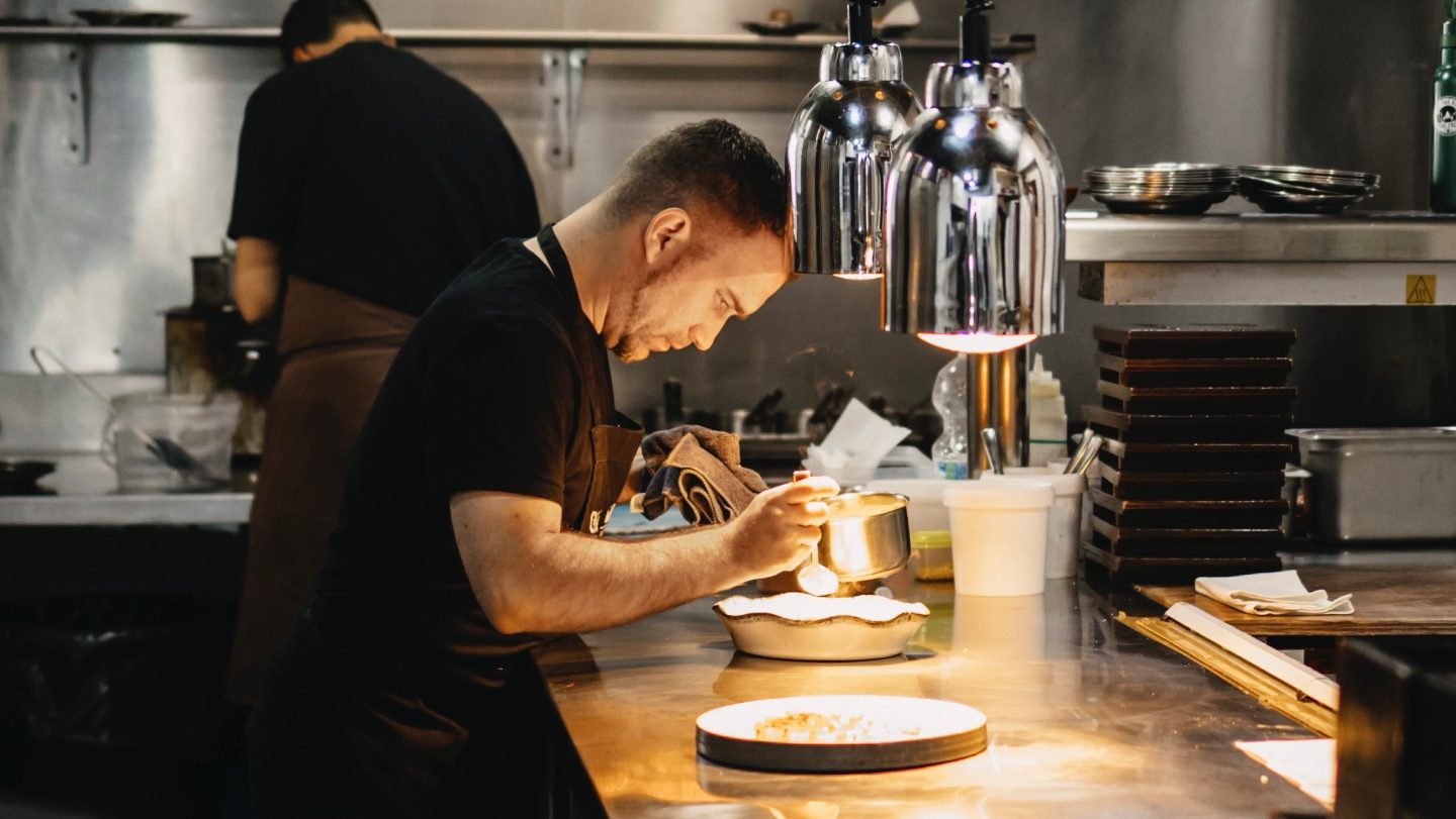 A focused chef plating a dish under warm kitchen lights, with another chef working in the background.