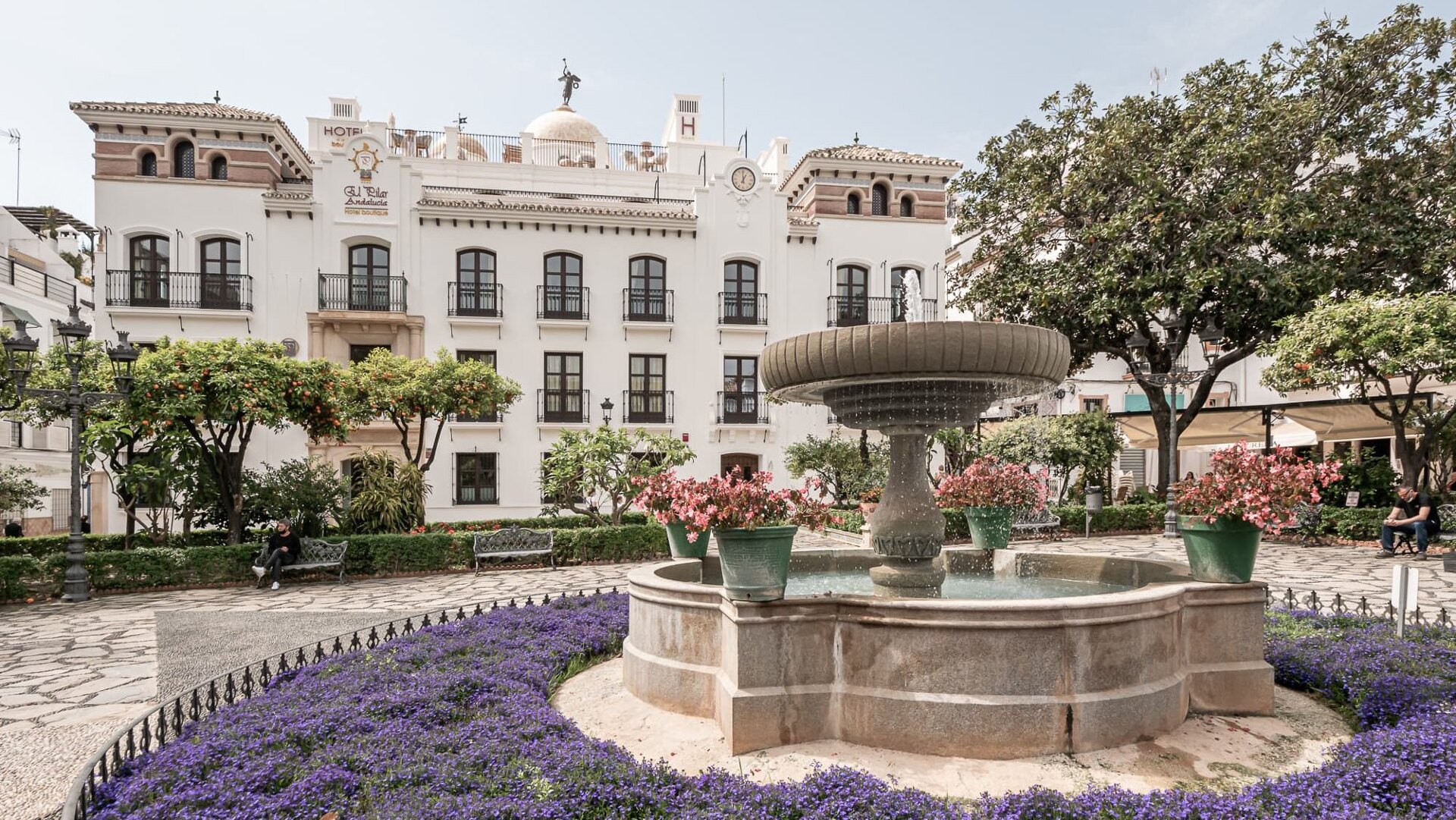 Picturesque square in Estepona Old Town, featuring a stone fountain surrounded by vibrant flowers, orange trees, and traditional Andalusian architecture in the background.