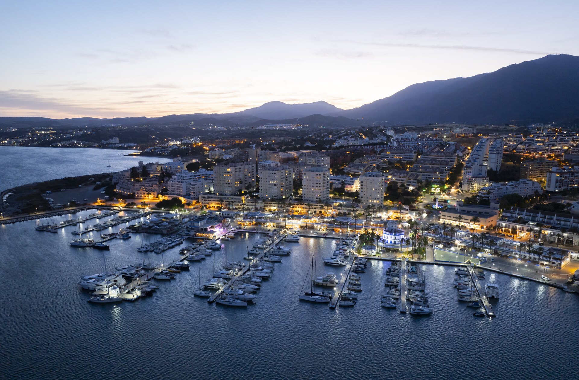 Aerial view of Estepona Marina at dusk, with yachts docked in the harbour, illuminated waterfront restaurants, and the cityscape extending towards the mountains.