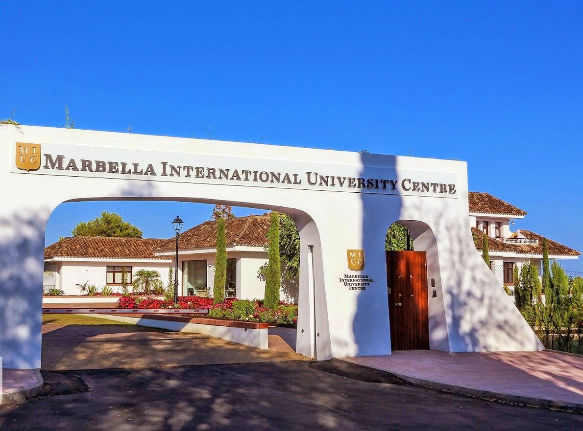 Entrance of Marbella International University Centre with a white archway, terracotta roofs, and lush greenery under a clear blue sky.