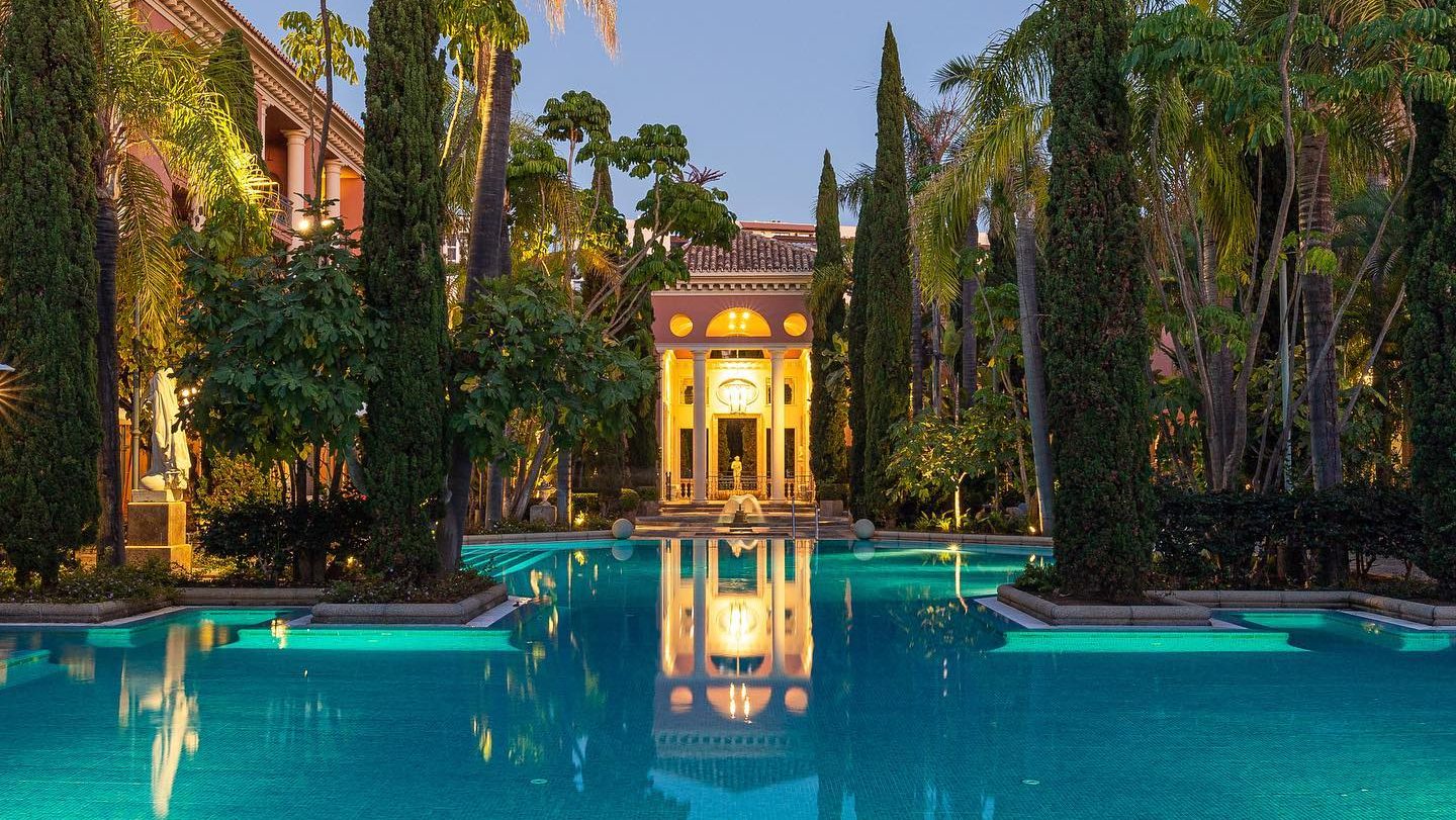 Illuminated neoclassical pavilion at Villa Padierna, reflected in the serene waters of a grand swimming pool, surrounded by lush Mediterranean gardens and tall cypress trees.