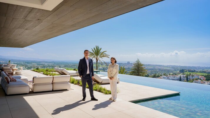 Founders Evelin and Frederic of Marbella Hills Homes standing on a luxury villa terrace in Marbella with infinity pool and panoramic sea views, representing a private and high-end real estate advisory on the Costa del Sol
