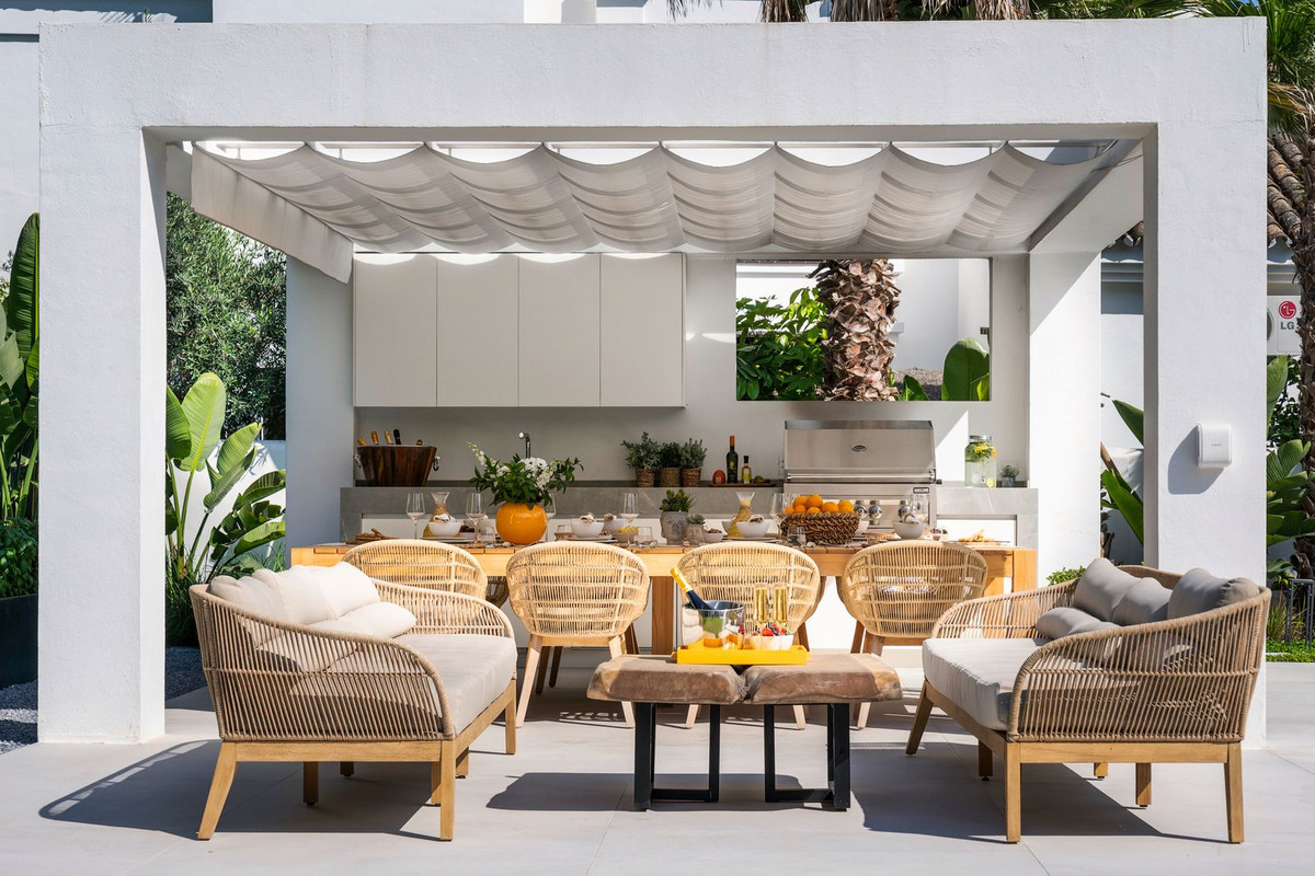 Close up of an outdoor kitchen and dining area under a white pergola, with wicker chairs and a sofa.