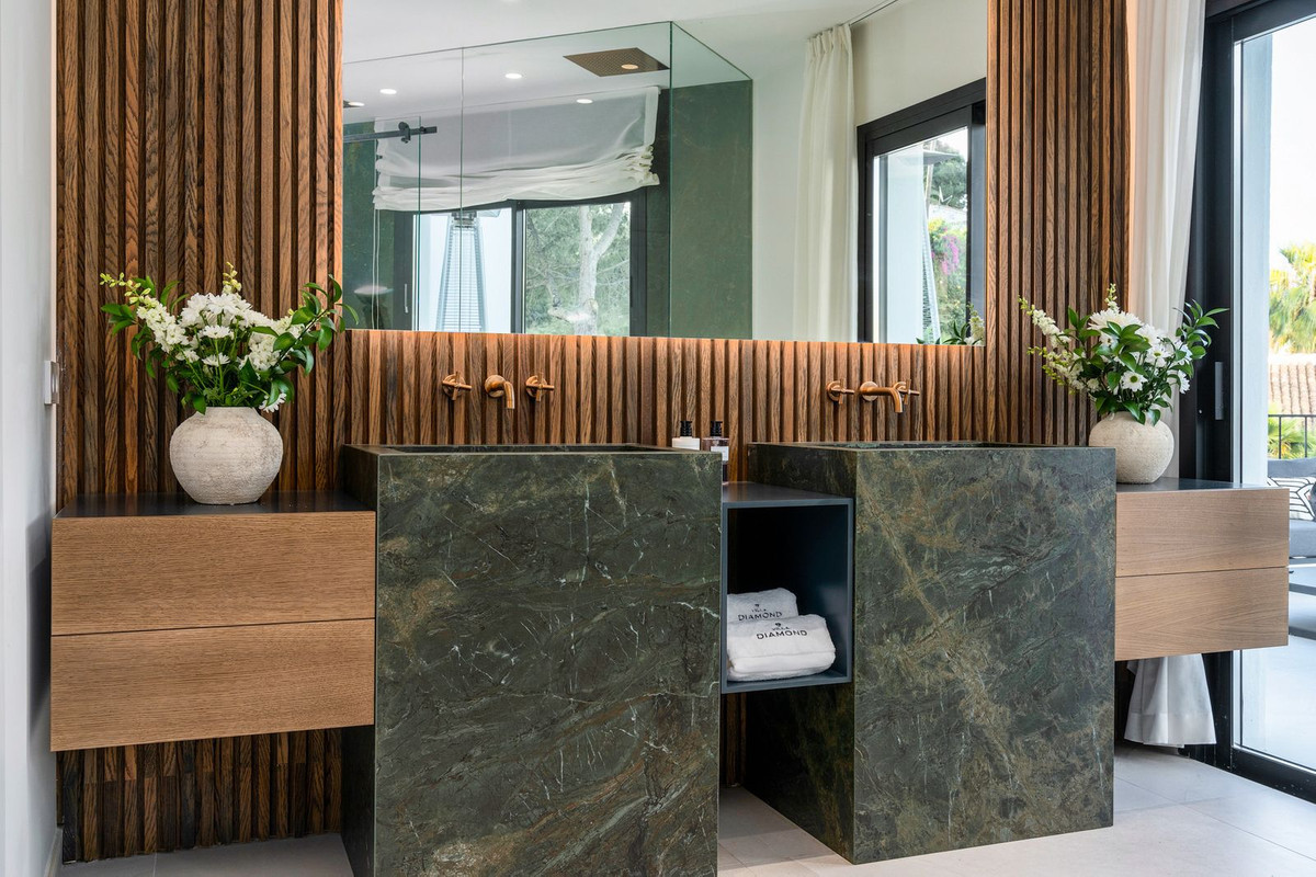 Bathroom with a wooden slatted wall, dual dark marble sinks, large mirror, and white flower arrangements.