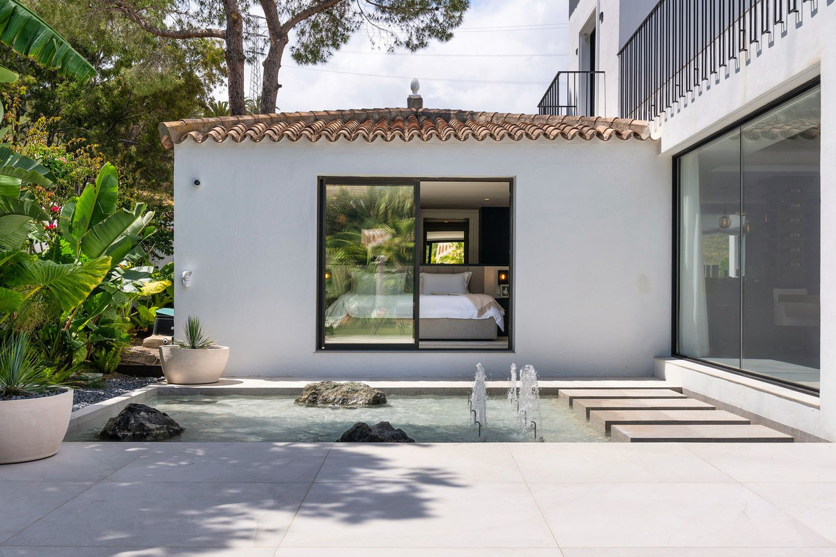 Exterior view of a guesthouse with a window looking into a bedroom, beside a decorative fountain and patio.