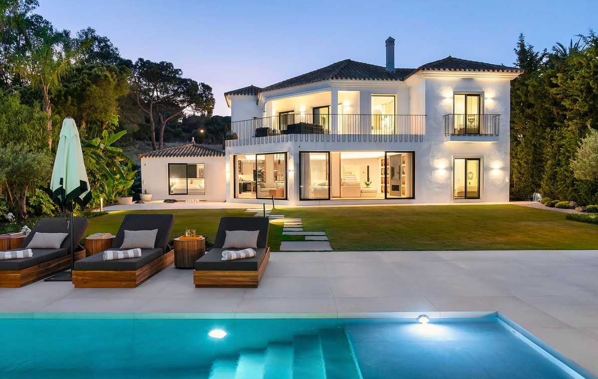 Exterior view of a white luxury villa with pool, sun loungers, and lush green garden at twilight.
