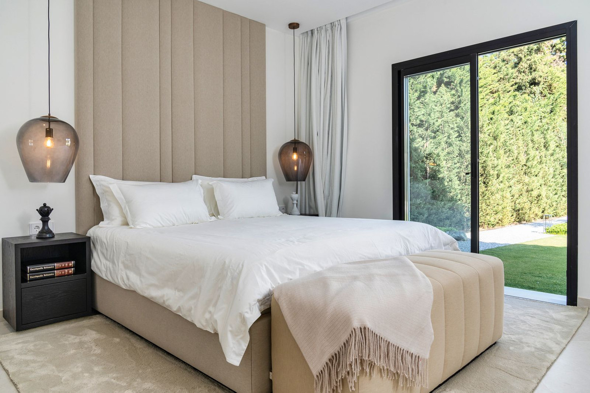 Bedroom with a large white bed, beige upholstered headboard, beige ottoman, and glass door to greenery.