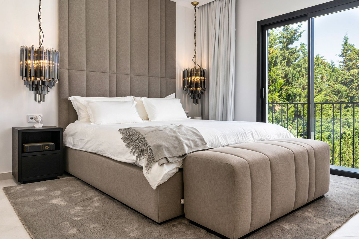 Bedroom with a large bed, upholstered headboard, hanging light fixtures, and a balcony with tree views.